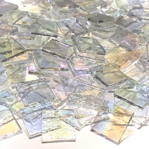 May include: A pile of iridescent glass mosaic tiles in various shades of light blue and green. The tiles are square shaped and have a smooth, glossy finish.