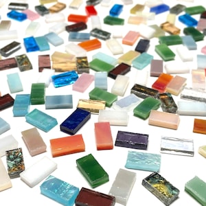 May include: A collection of small, rectangular glass tiles in various colors and patterns. The tiles are scattered on a white surface.