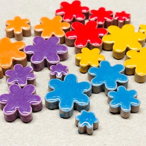 May include: A collection of small, six-petaled flower-shaped ceramic tiles in various glossy colors including red, orange, yellow, purple, and blue. Each piece features a smooth, reflective surface and a matte, unglazed base visible on the sides.
