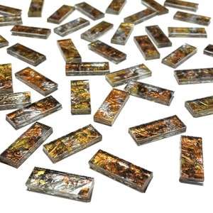 May include: A collection of small, rectangular glass tiles with a metallic gold and silver finish. The tiles have a textured, iridescent surface and are scattered on a white background.