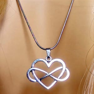 May include: Silver necklace with a pendant that merges a heart and an infinity symbol. The pendant is suspended from a fine silver chain. The necklace is shown against a plain backdrop.