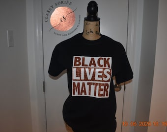 May include: Black Lives Matter t-shirt with the text printed in white letters on a brown glitter background.