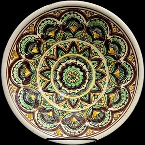 May include: A decorative ceramic plate with a detailed floral pattern. The design features a central flower surrounded by concentric rings of stylized leaves and floral motifs in shades of green, brown, and yellow against a cream background.