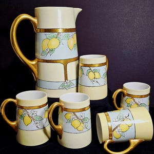 Könnte beinhalten: Ein Set aus drei Keramikbechern mit gesprenkelter, cremefarbener Glasur. Jeder Becher hat ein handgemaltes Design: ein blau-grünes Blatt, eine gelbe Blume und eine rote Beere. Die Becher haben ein schlichtes, modernes Design.