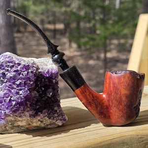 May include: A wooden tobacco pipe with a curved stem and a dark finish, resting next to a large, purple amethyst geode. The pipe has a reddish-brown bowl and a black mouthpiece. The geode is a cluster of purple crystals.