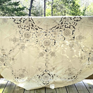 Puede incluir: Mantel redondo color crema con intrincados bordados florales y geométricos. El bordado presenta un color marrón oscuro, creando un contraste llamativo con el fondo más claro. El mantel está drapeado sobre una barandilla de madera.