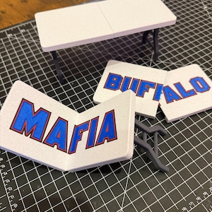 May include: A miniature white folding table with dark gray legs and three separate white rectangular pieces. Two pieces feature the blue and red text BUFFALO, and one folded piece features the blue and red text MAFIA. The items are displayed on a black cutting mat.