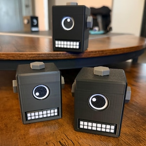 May include: Three black, box-shaped robot figures with a single large circular eye, a wide rectangular mouth filled with white square teeth, and a small gray hexagonal knob on top. Each robot has small gray side protrusions and a smooth finish.