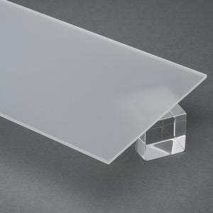 May include: A rectangular, frosted glass panel rests on a clear, faceted cube. The glass has a smooth, matte finish and is angled, showcasing its translucent quality. The background is a solid, dark gray.