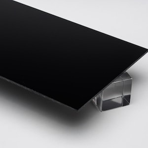 May include: A black rectangular object, possibly a panel or sheet, is angled slightly upwards, resting on a clear, cube-shaped support. The surface is smooth and reflects light, creating a contrast against the white background.