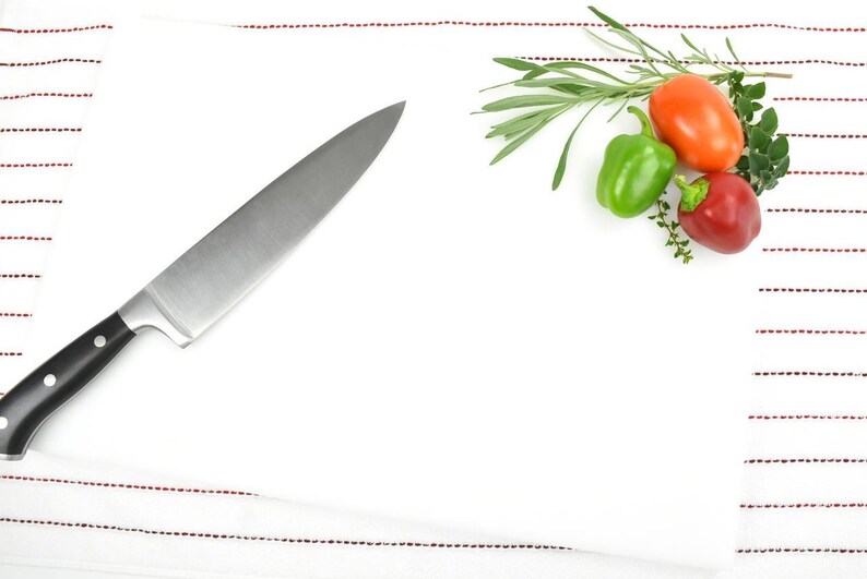 May include: A white kitchen towel with red stripes, a chef's knife, and a sprig of rosemary with red and green bell peppers.