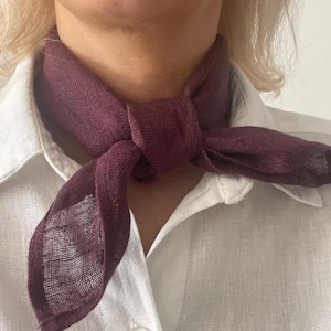 May include: A burgundy scarf tied in a knot, worn over a white button-down shirt. The scarf is made of a lightweight, textured fabric. The image is a close-up of the neck and upper chest.