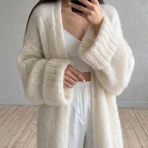 Könnte beinhalten: Cremefarbene, lange Strickjacke mit offenem Schnitt und weiten Ärmeln. Das Strickwerk wirkt weich und flauschig. Die Strickjacke wird über einem weißen Oberteil und einer weißen Hose getragen. Die Ärmel haben gerippte Bündchen. Das Outfit hat eine neutrale Farbpalette.