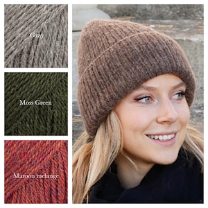 May include: A brown knitted beanie hat is worn by a person with blonde hair. The image also shows three skeins of yarn in grey, moss green, and maroon melange colours. The yarn is likely for knitting or crocheting projects.