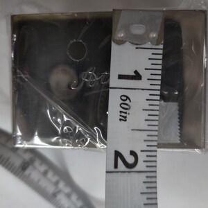 May include: A silver metal plate with a round hole and the word "Paris" engraved on it. The plate is being measured with a tape measure that reads 1 60/in.