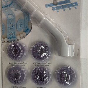 Puede incluir: Un juego de punzones de fondant morado con un mango de plástico blanco. Los punzones incluyen un diamante estrecho con volutas, un diamante ancho con volutas, un tulipán grande con hojas, una flor holandesa, un paisley con puntos, un tulipán pequeño con hojas, un boca de dragón con hojas y un trébol de cuatro hojas con puntos. El embalaje incluye el texto "Fondant Decorative Punch Set" y "Ensemble de poinçons pour le fondant".