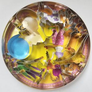 Könnte beinhalten: Ein Satz Metall-Ausstecher in einer runden Dose. Die Ausstecher sind in Form verschiedener Tiere und Gegenstände, darunter ein Ballon, ein Mädchen, eine Kuh und ein Pferd. Die Dose hat einen gelben Hintergrund mit rosa und weißen Streifen.