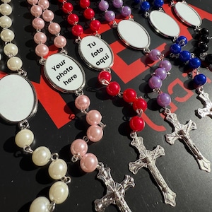 Op de afbeelding: Een verzameling rozenkransen in diverse kleuren, waaronder wit, roze, rood, paars, blauw en zwart. Elke rozenkrans heeft een zilveren kruis en een ovale hanger met de tekst "Your photo here" of "Tu foto aquí".