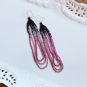 May include: A pair of long, beaded earrings with a gradient of colors from black to purple to pink. The earrings are made with small, round beads and have silver hooks.
