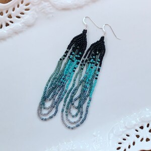 May include: A pair of long, beaded earrings with a gradient of colors from black to turquoise. The earrings are made with small, seed beads and have silver hooks.