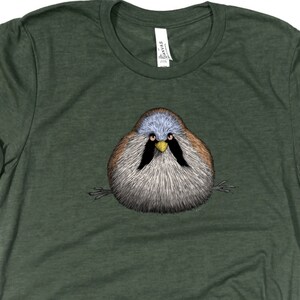Könnte beinhalten: Ein dunkelgrünes T-Shirt mit einer Cartoon-Illustration eines runden, flauschigen Vogels mit einem schwarzen Schnabel und einem gelben Streifen über seinem Schnabel. Der Vogel hat einen blauen Kopf und einen braunen Körper.