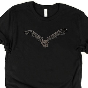 K&ouml;nnte beinhalten: Schwarzes T-Shirt mit einer detaillierten Illustration einer Fledermaus im Flug. Die Fledermaus ist mit ausgebreiteten Fl&uuml;geln dargestellt, die filigrane Linienarbeit zeigen. Das Design ist auf dem T-Shirt zentriert und bietet einen auff&auml;lligen Anblick auf dem dunklen Stoff. Das T-Shirt hat hochgekrempelte &Auml;rmel.