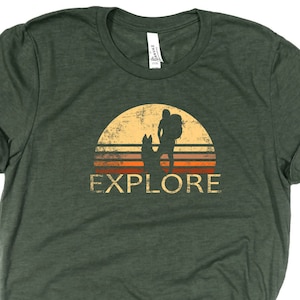 May include: Green t-shirt with a distressed graphic of a hiker with a dog silhouetted against a sunset. The word "EXPLORE" is printed below the image in a vintage style.