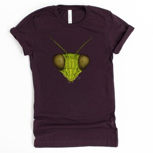 May include: A dark purple t-shirt with a green praying mantis face graphic printed on the front. The mantis has large, round eyes and a detailed illustration.