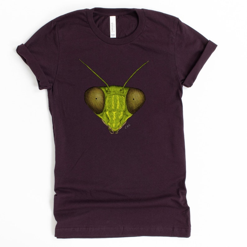 May include: A dark purple t-shirt with a green praying mantis face graphic printed on the front. The mantis has large, round eyes and a detailed illustration.