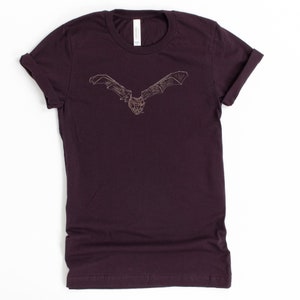 K&ouml;nnte beinhalten: Ein dunkelburgunderfarbenes T-Shirt mit einer detaillierten Illustration einer Fledermaus mit ausgebreiteten Fl&uuml;geln. Die Fledermaus ist in einem hellbraunen Farbton dargestellt, der einen Kontrast zum tiefen Farbton des Shirts bildet. Das Shirt hat hochgekrempelte &Auml;rmel und einen Rundhalsausschnitt.