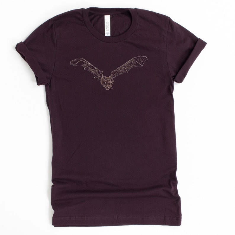 K&ouml;nnte beinhalten: Ein dunkelburgunderfarbenes T-Shirt mit einer detaillierten Illustration einer Fledermaus mit ausgebreiteten Fl&uuml;geln. Die Fledermaus ist in einem hellbraunen Farbton dargestellt, der einen Kontrast zum tiefen Farbton des Shirts bildet. Das Shirt hat hochgekrempelte &Auml;rmel und einen Rundhalsausschnitt.