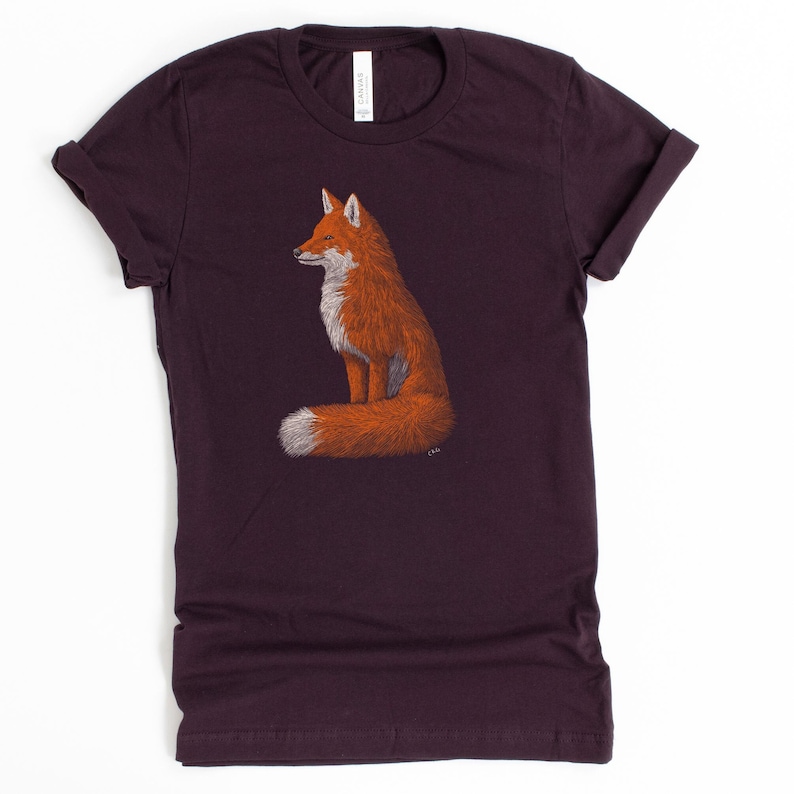 May include: A dark purple t-shirt featuring a graphic of a red fox sitting with its tail curled around its body.