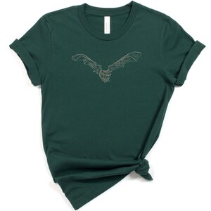 K&ouml;nnte beinhalten: Dunkelgr&uuml;nes T-Shirt mit einer detaillierten, hellfarbigen Illustration einer Fledermaus mit ausgebreiteten Fl&uuml;geln. Das Shirt besteht aus einem weichen Material und hat hochgekrempelte &Auml;rmel. Das Shirt ist unten geknotet.
