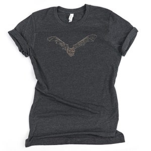 K&ouml;nnte beinhalten: Dunkelgraues T-Shirt mit einer detaillierten Illustration einer Fledermaus mit ausgebreiteten Fl&uuml;geln. Das Fledermaus-Design ist hellbraun und mittig auf der Vorderseite platziert. Das Shirt hat hochgekrempelte &Auml;rmel und einen Rundhalsausschnitt.