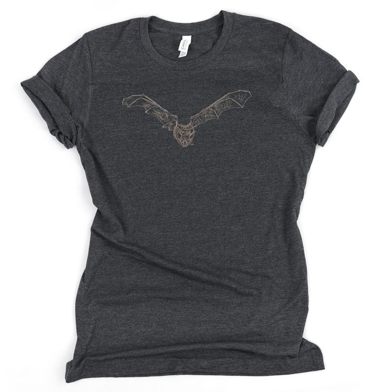 K&ouml;nnte beinhalten: Dunkelgraues T-Shirt mit einer detaillierten Illustration einer Fledermaus mit ausgebreiteten Fl&uuml;geln. Das Fledermaus-Design ist hellbraun und mittig auf der Vorderseite platziert. Das Shirt hat hochgekrempelte &Auml;rmel und einen Rundhalsausschnitt.