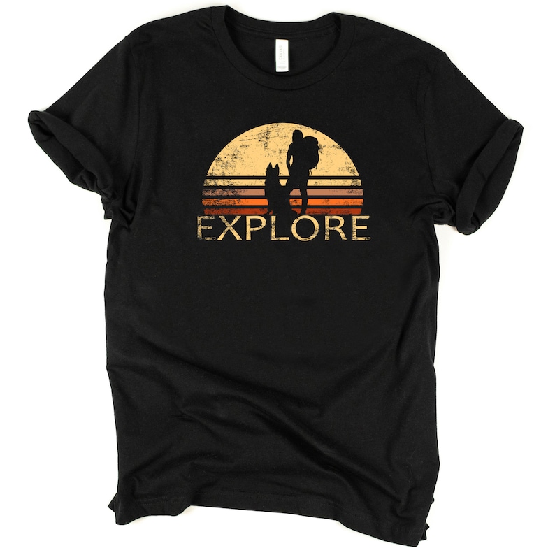 May include: Black t-shirt with a retro design featuring a hiker with a dog silhouetted against a sunset. The word "EXPLORE" is printed in a distressed font below the image. A comfortable tee for outdoor enthusiasts.