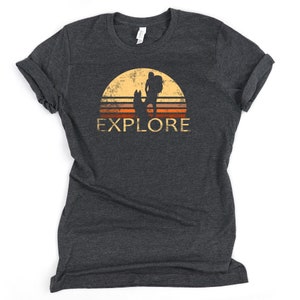 May include: Dark gray t-shirt with a retro design featuring a hiker with a dog silhouetted against a sunset. The word "EXPLORE" is printed in a distressed font below the image. The shirt has rolled-up sleeves.
