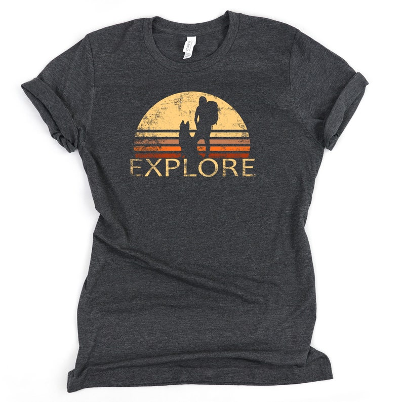 May include: Dark gray t-shirt with a retro design featuring a hiker with a dog silhouetted against a sunset. The word "EXPLORE" is printed in a distressed font below the image. The shirt has rolled-up sleeves.