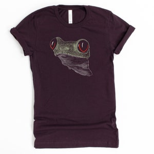 May include: A dark purple t-shirt with a white and green frog illustration. The frog has large red eyes.