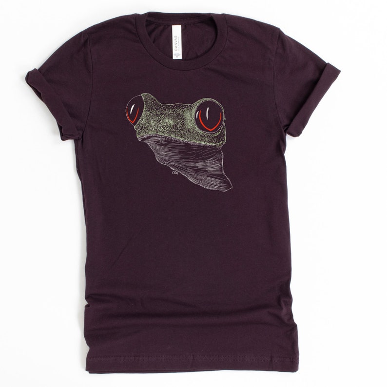 May include: A dark purple t-shirt with a white and green frog illustration. The frog has large red eyes.