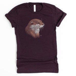 May include: A dark purple t-shirt with an embroidered image of an otter's head. The otter is facing right and has brown fur with a white muzzle.