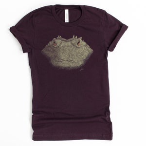 May include: A maroon t-shirt featuring a detailed illustration of a reptile's face. The artwork is in shades of gray and tan, with red eyes and a textured appearance. The shirt has rolled-up sleeves and a crew neck.