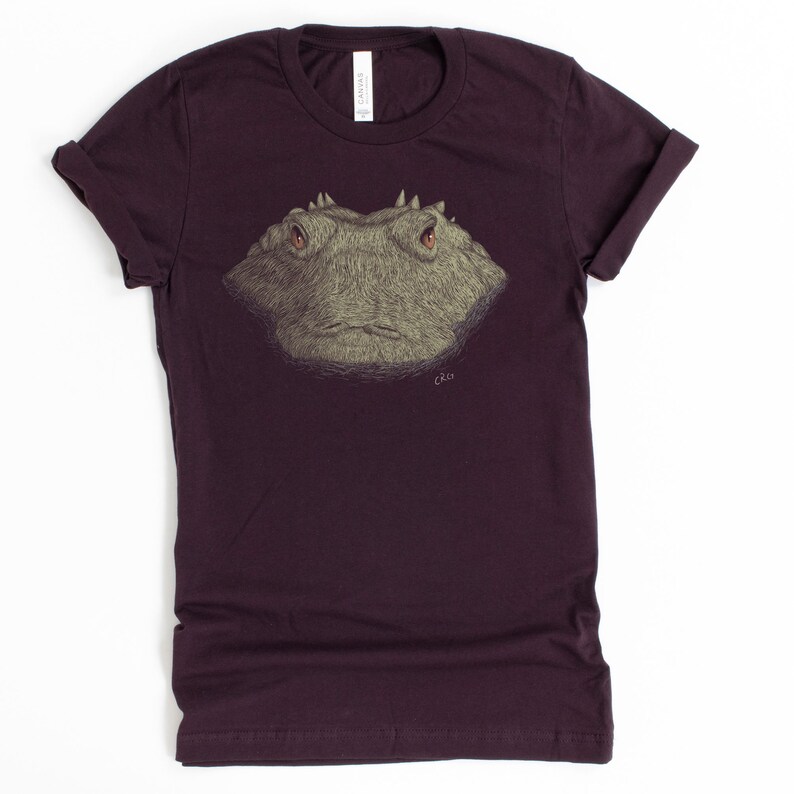 May include: A maroon t-shirt featuring a detailed illustration of a reptile's face. The artwork is in shades of gray and tan, with red eyes and a textured appearance. The shirt has rolled-up sleeves and a crew neck.