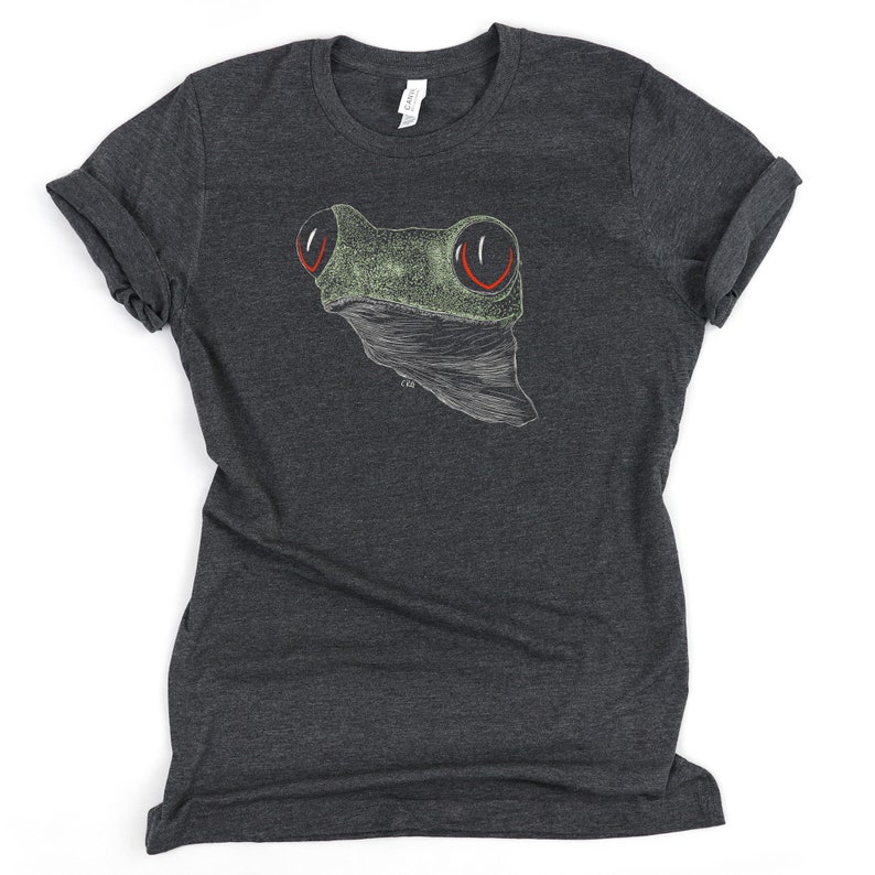 May include: A dark heather grey t-shirt with a white outline drawing of a frog with red eyes. The frog is facing the viewer.
