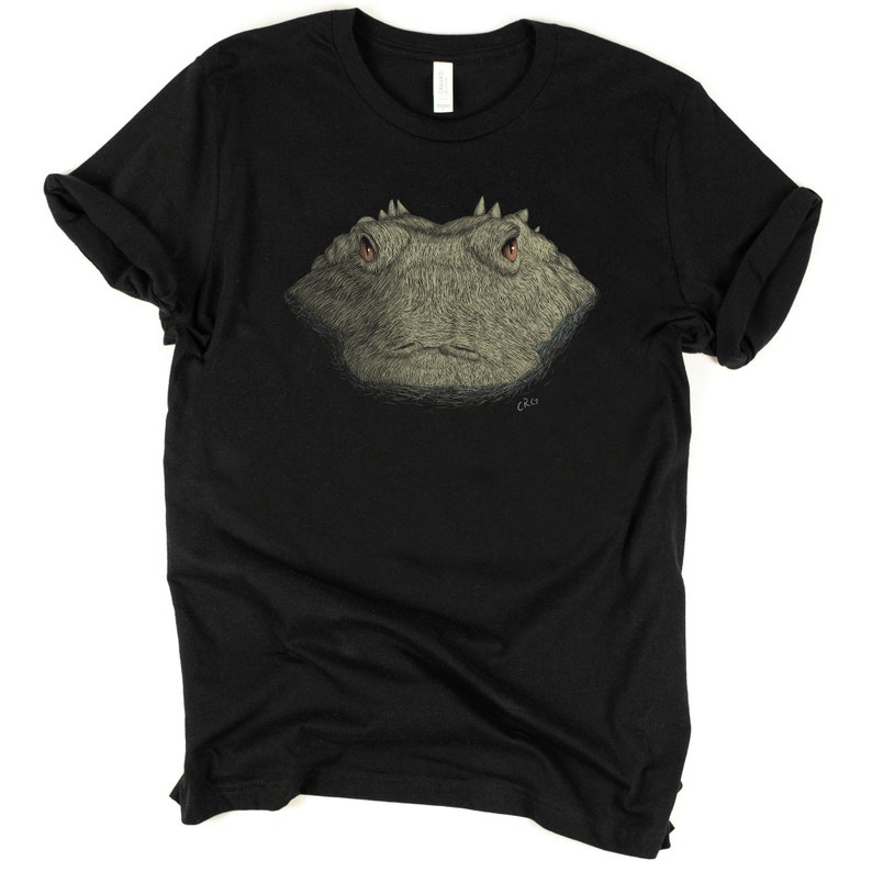 May include: Black t-shirt featuring a detailed illustration of a reptile's face. The reptile has textured, gray skin, red eyes, and a serious expression. The artist's signature is in the lower right corner. The shirt is made of a soft material.