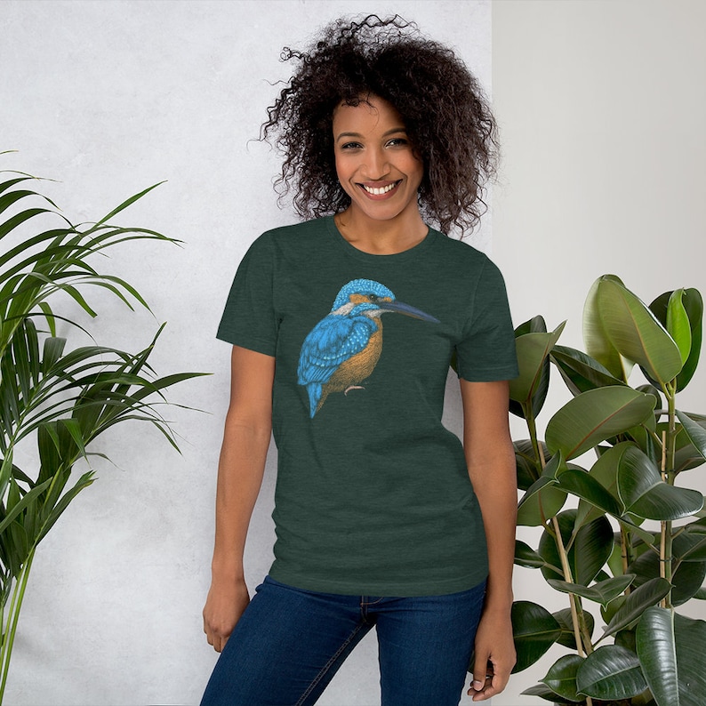 May include: A woman wearing a dark green t-shirt with a blue and brown kingfisher bird graphic. The bird is facing left and has a long, thin beak.