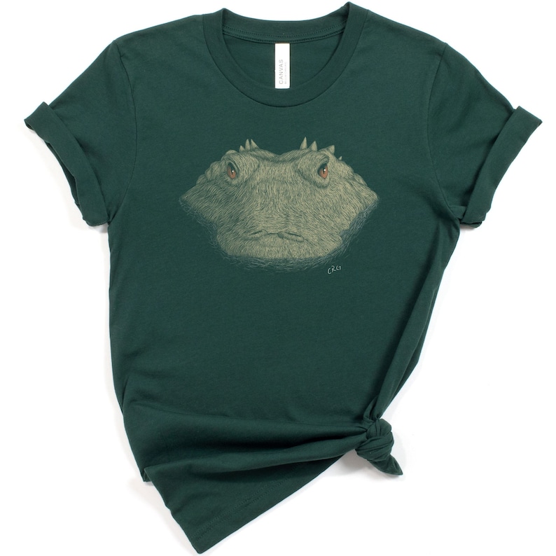 May include: A dark green t-shirt featuring a detailed illustration of a frog's face. The frog has textured skin, red eyes, and a serious expression. The shirt is made of a soft material and has rolled-up sleeves. The brand name "CANVAS" is printed on the tag.