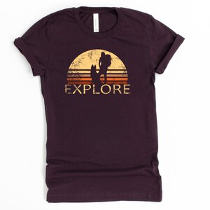 May include: A dark maroon t-shirt with a distressed graphic of a hiker and dog silhouetted against a sunset. The word "EXPLORE" is printed below the image in a vintage font. The shirt is made of a soft material.