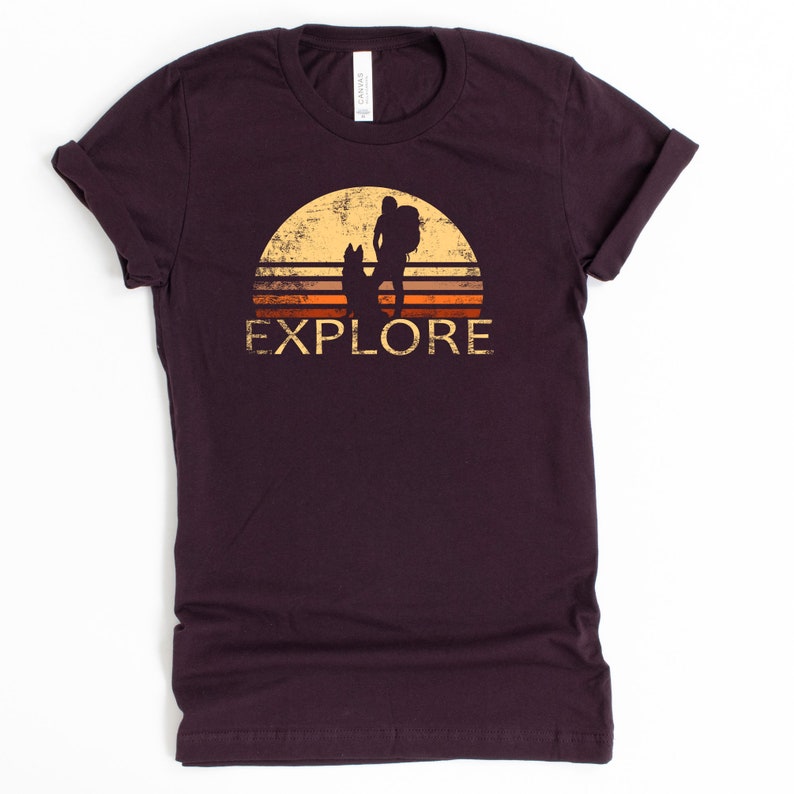 May include: A dark maroon t-shirt with a distressed graphic of a hiker and dog silhouetted against a sunset. The word "EXPLORE" is printed below the image in a vintage font. The shirt is made of a soft material.