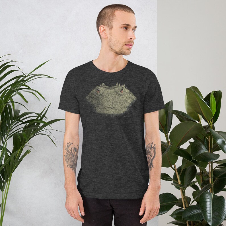 May include: Dark gray t-shirt featuring a detailed illustration of a frog's face. The frog's face is in shades of green and brown, with visible eyes and textured skin. The t-shirt is made of a soft material and has short sleeves.